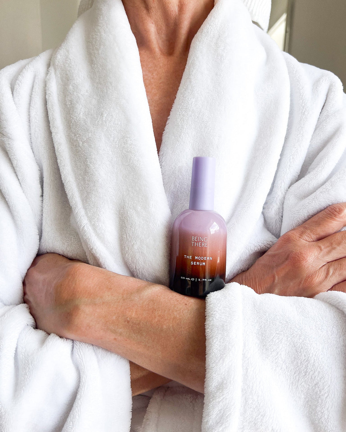 A person wearing a white bathrobe holds a bottle of peptide serum. The bottle is a gradient of lavender purple to dark brown, with white text that reads THE MODERN SERUM.